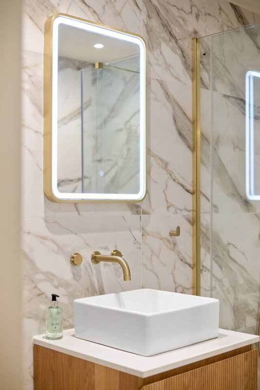 Modern bathroom with marble walls, illuminated mirror, white vessel sink and brass fixtures