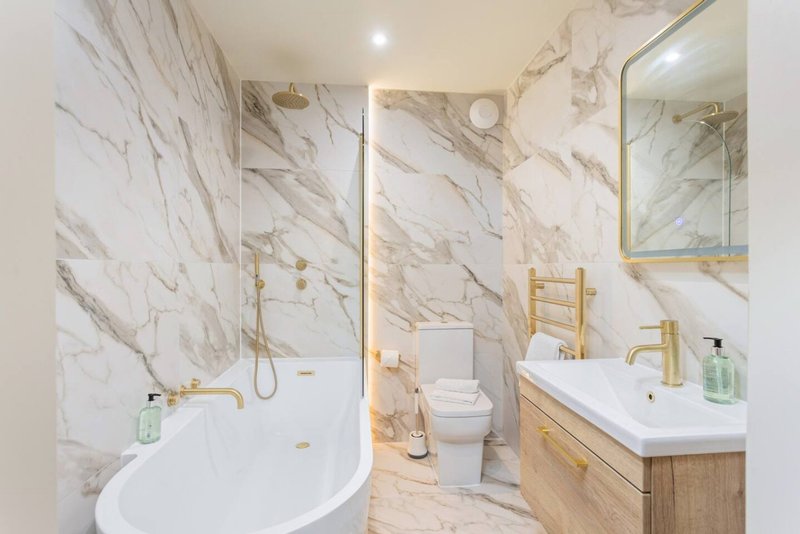 Modern bathroom with marble walls, curved white bathtub, wooden vanity and gold fixtures
