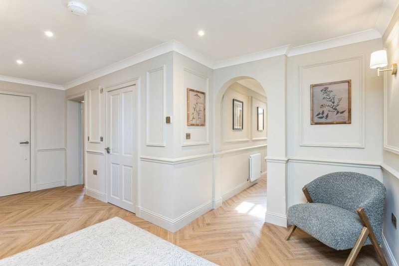 Bright hallway with white paneled walls, wooden flooring, framed artwork, and modern armchair