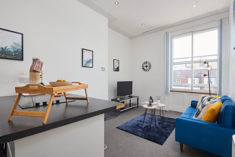 Modern living room with blue sofa, wooden desk, TV unit, and large windows overlooking street