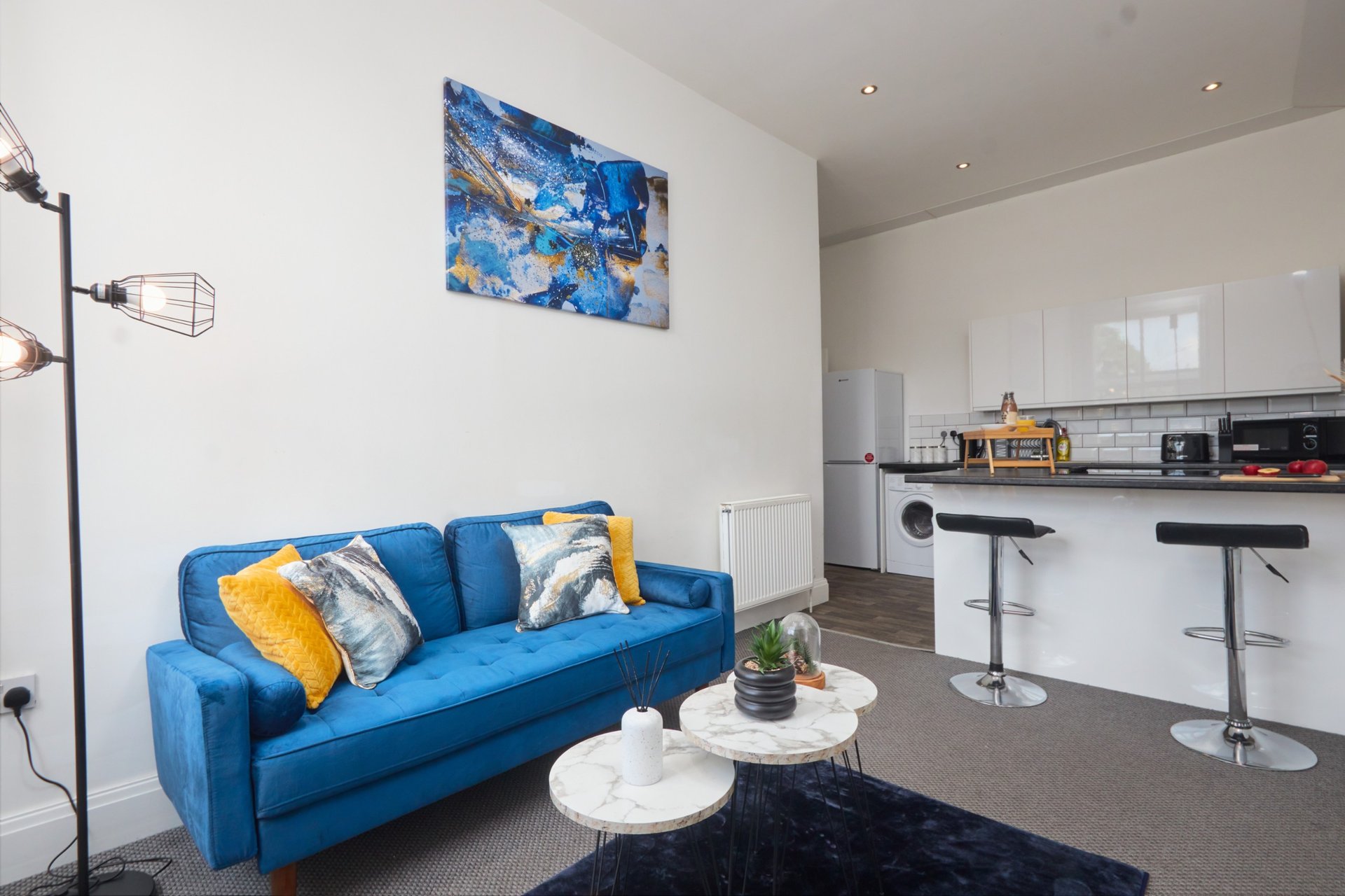 Open-plan living area with blue sofa, white kitchen, bar seating, and abstract artwork