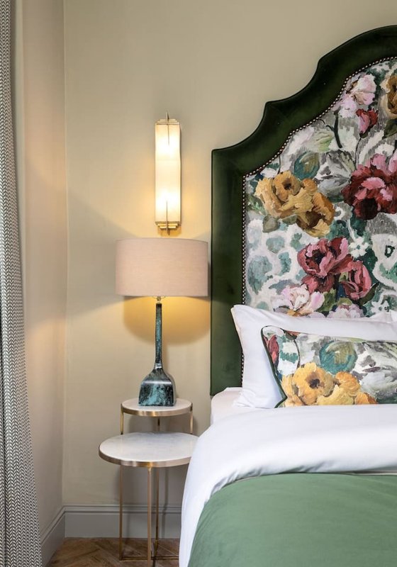 Bedroom corner with green upholstered headboard, floral patterned pillows, bedside lamp and small table