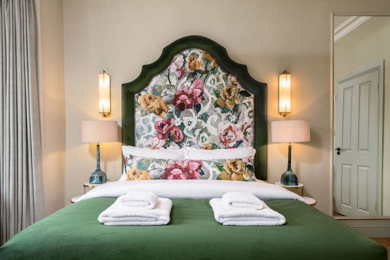 Bedroom with floral upholstered headboard, green bedspread, folded towels, and bedside lamps