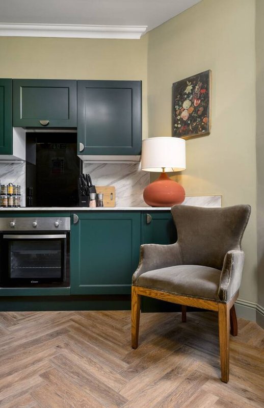 Kitchen and living area with green cabinetry, upholstered chair, lamp, and floral wall art