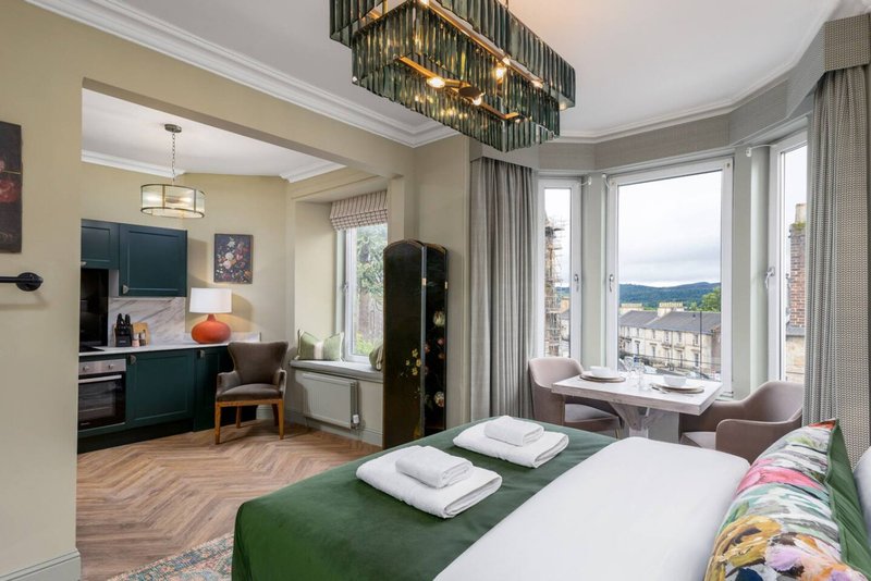 Modern bedroom with double bed, green throw, white pillows, city views through large windows and open-plan kitchen area