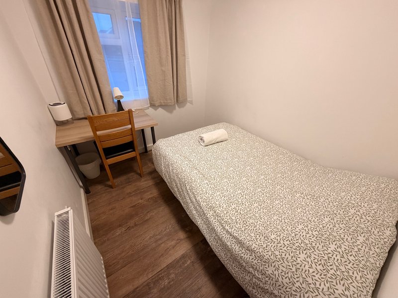 Small bedroom with single bed, wooden desk, chair, and window with beige curtains