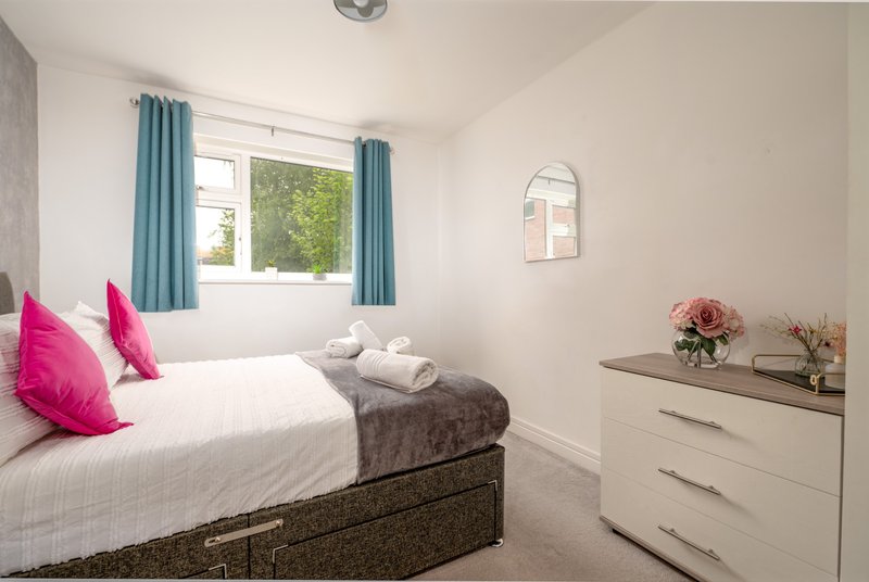 Bright bedroom with double bed, pink cushions, teal curtains, and wooden dresser
