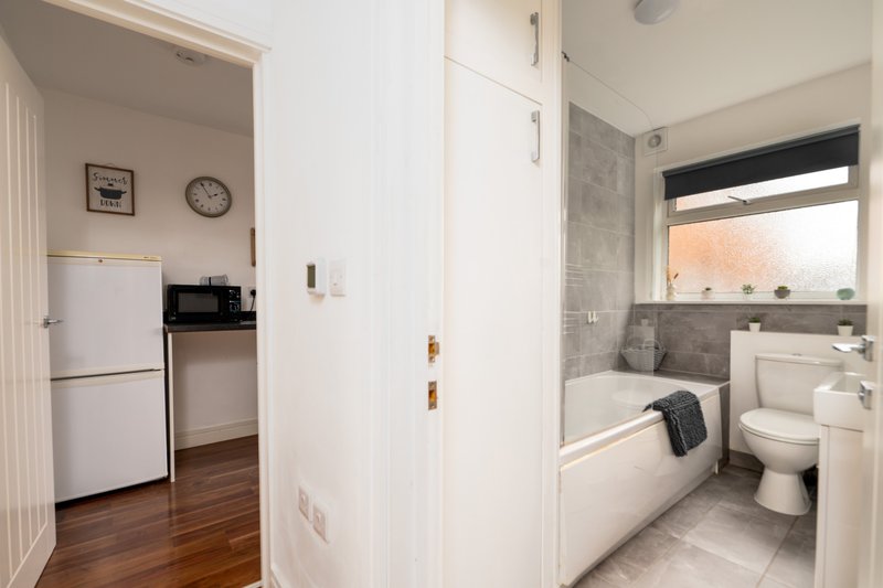 Modern bathroom with white suite, grey tile walls, and window; kitchen with white fridge visible through doorway