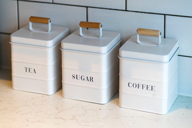 Three white kitchen storage canisters with wooden handles labeled tea, sugar, and coffee