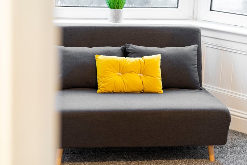 Modern grey sofa bed with yellow cushion, wooden legs, and window behind