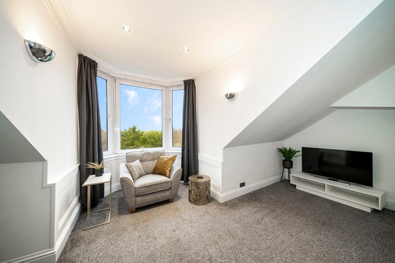 Modern attic bedroom with angled ceilings, armchair by bay window, television, and carpet flooring