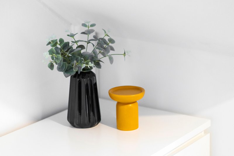 Minimalist shelf display with black vase of green foliage and yellow ceramic stool