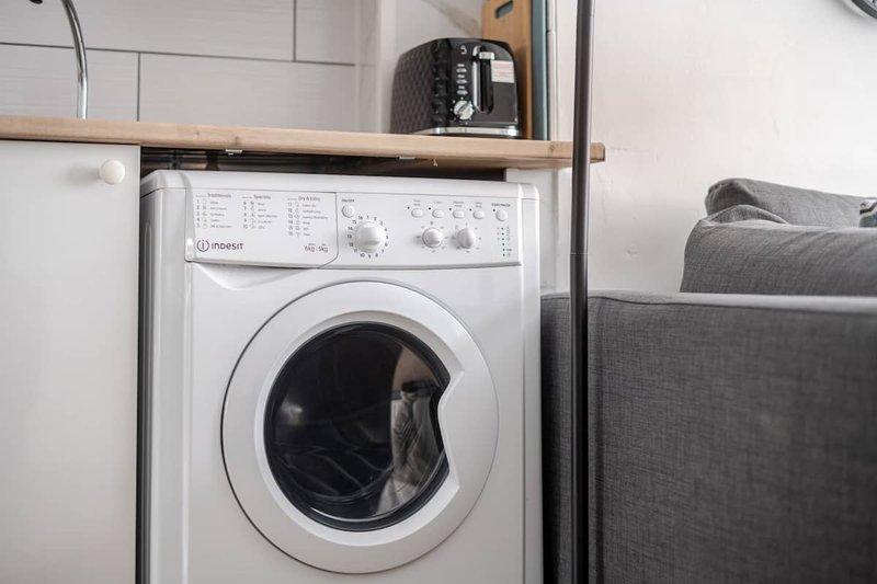 Laundry area with white front-load washing machine, wooden shelf above, and gray upholstered furniture