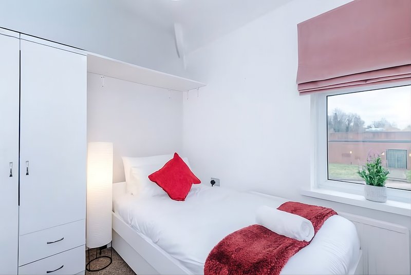 Bright bedroom with single bed, red cushions, wardrobe and window with pink blind and also storage space