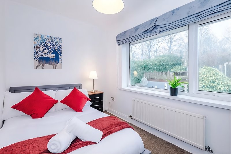 Bright bedroom with double bed, red cushions, white bedding and large window with garden view and storage space