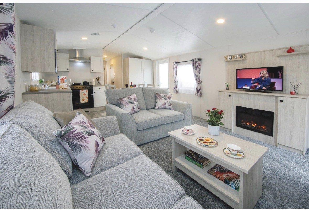 Open-plan living area with gray sofas, fireplace, kitchen, and dining space in modern caravan