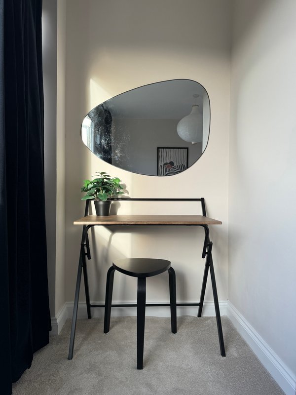 Minimalist vanity area with organic mirror, wooden desk, black stool and potted plant