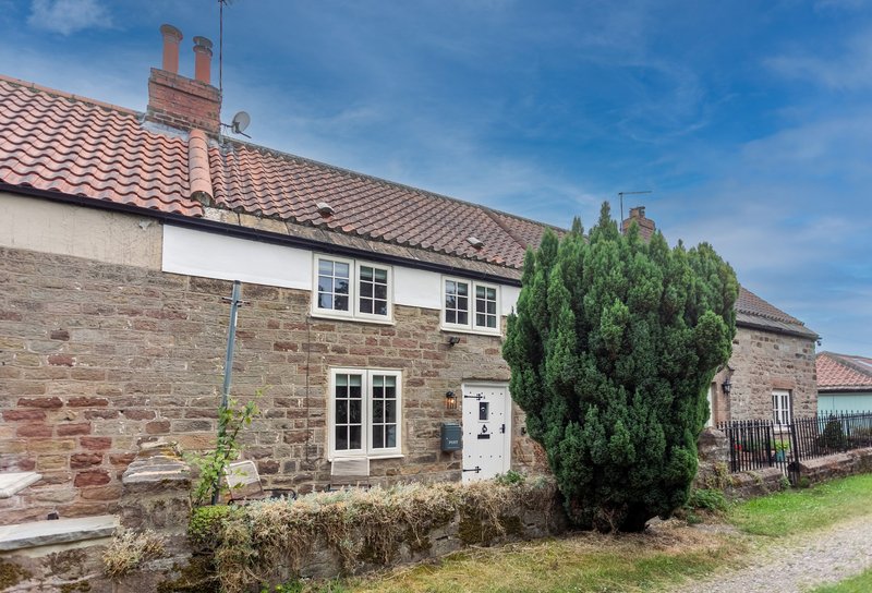 Stone and brick cottage with tiled roof, white door and windows, surrounded by garden