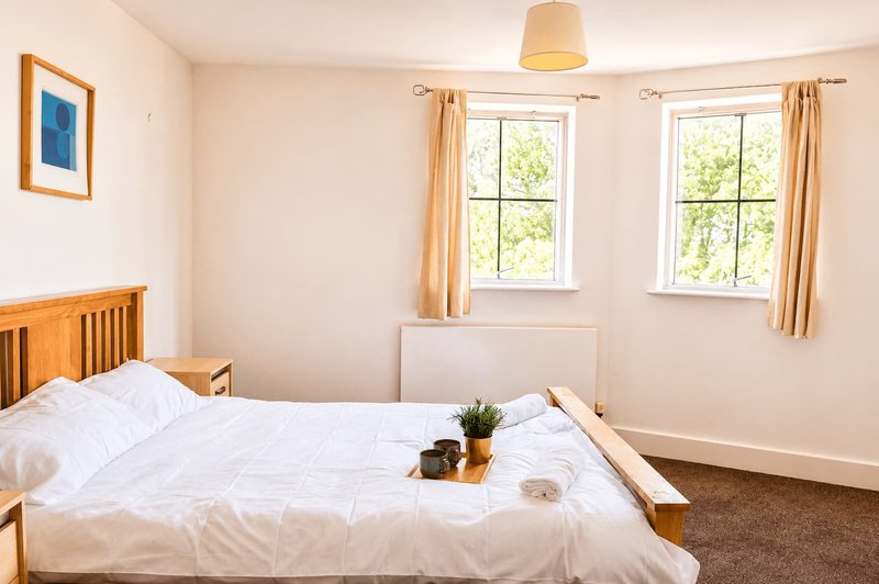 Bright bedroom with double bed, wooden frame, two windows with cream curtains, and pendant light