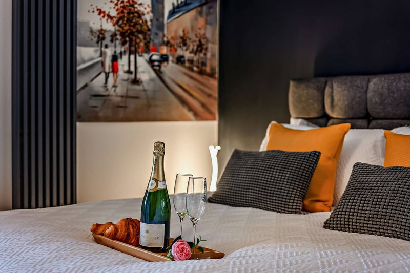 Bedroom with champagne bottle, glasses, pastries on bed, orange and black cushions, framed artwork above