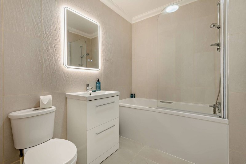 Modern bathroom with toilet, white vanity, illuminated mirror, and bathtub with glass partition