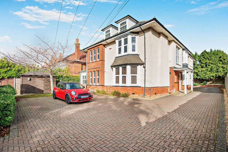 Modern residential property exterior with red car parked on brick driveway, manicured gardens, and clear blue sky