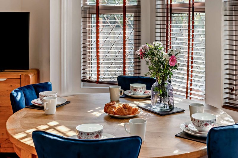 Dining room with round wooden table, blue upholstered chairs, fresh flowers, and shuttered windows