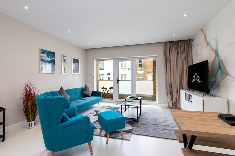 Modern living room with turquoise sofa and armchair, wooden desk, wall-mounted TV, and glass doors to balcony