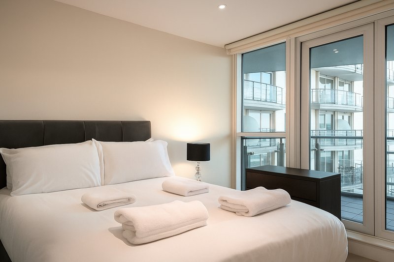 Modern bedroom with double bed, white bedding, black headboard, and balcony doors with city views