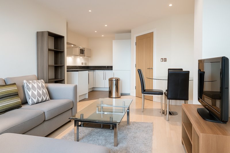 Open-plan living area with grey sofa, glass coffee table, TV stand and kitchenette
