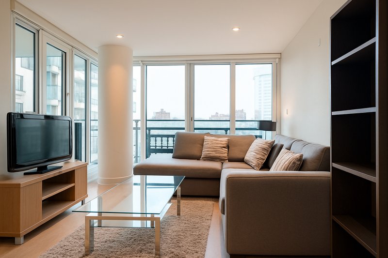 Modern living room with brown sectional sofa, TV on wooden stand, glass coffee table, and city views from floor-to-ceiling windows