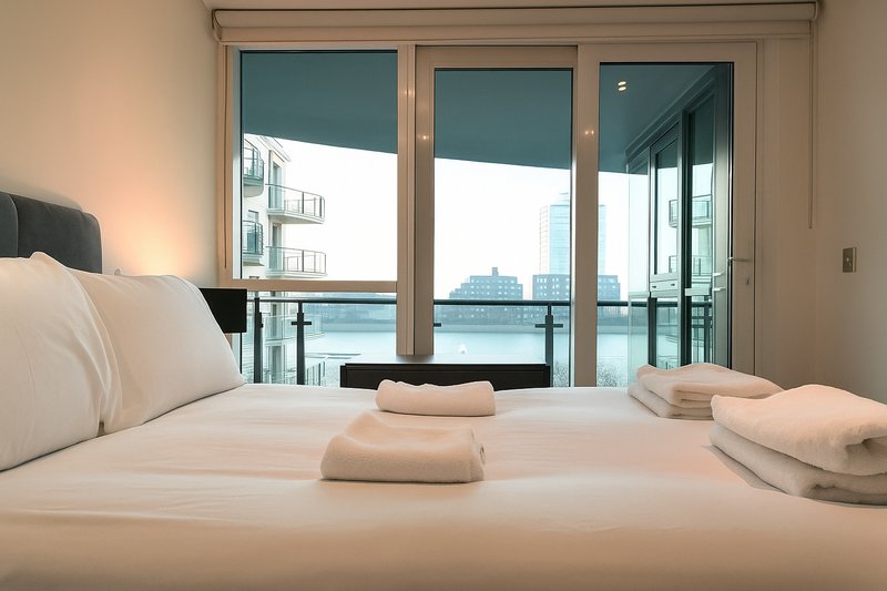 Modern bedroom with double bed, rolled towels, and floor-to-ceiling windows overlooking waterfront