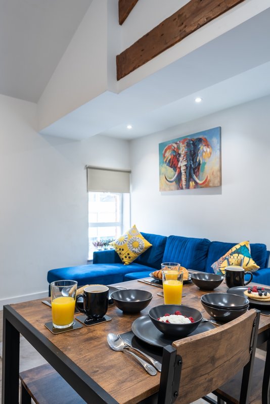 Open-plan living space with blue sofa, wooden beams, dining table set with breakfast items and drinks