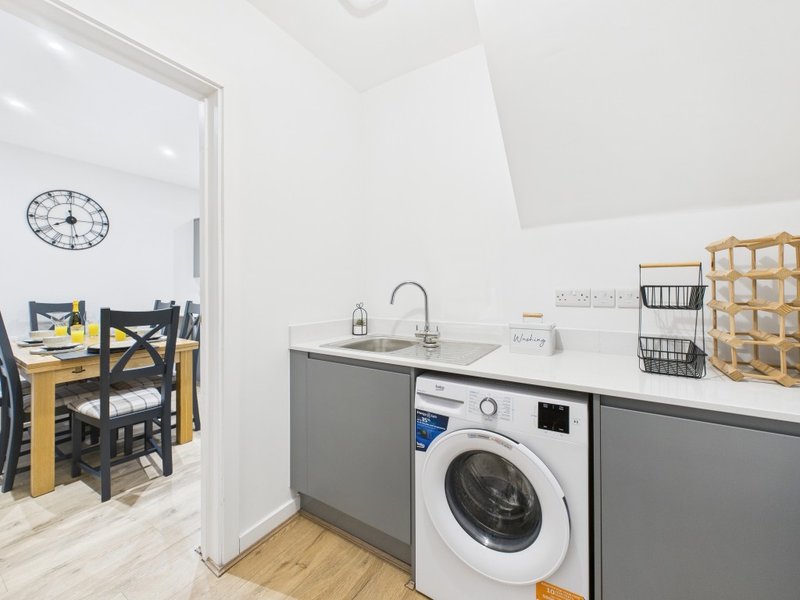 Modern laundry room with washing machine, gray cabinetry, sink, and wooden dining area visible through doorway