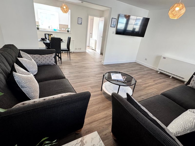 Open-plan living room with black sofas, wooden flooring, TV, and kitchen area visible
