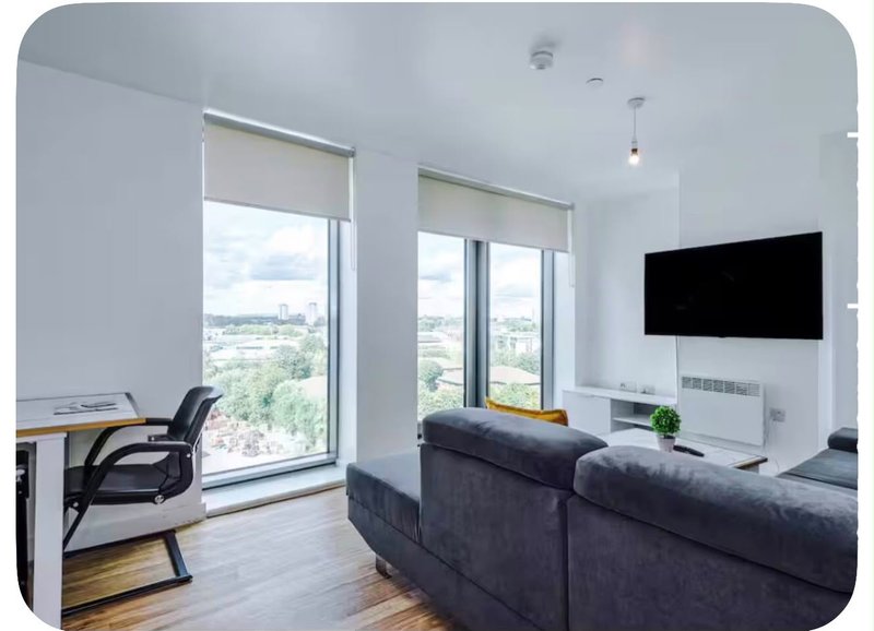 Modern living room with dark sofa, wall-mounted TV, desk with chair, and large windows with city views