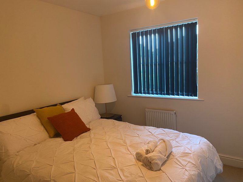 Bedroom with double bed, white bedding, rust cushion, bedside lamp and black blinds
