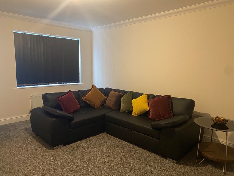 Living room with black corner sofa, colorful cushions, window with blinds, mounted smart TV and side table