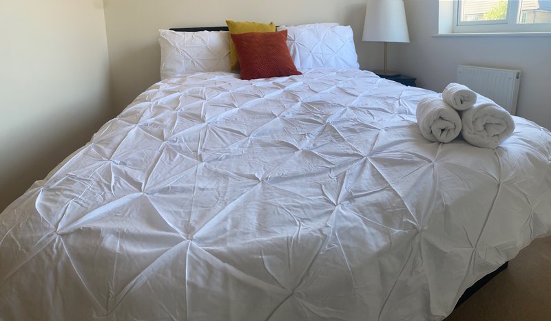 Bedroom with white pinched duvet, decorative pillows, rolled towels and window
