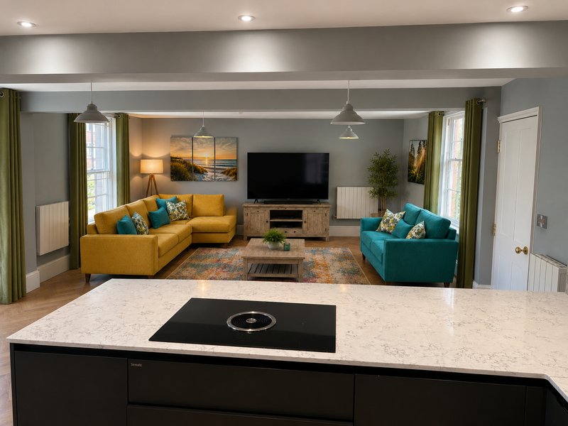 Open-plan living room with yellow and teal sofas, TV, kitchen island, and natural light from windows