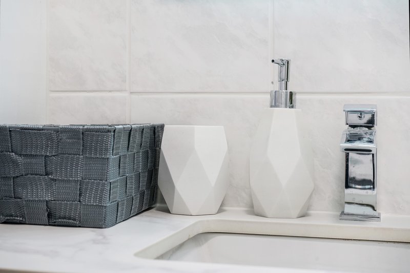 Bathroom counter with white ceramic accessories, soap dispenser, woven storage basket, and chrome fixtures