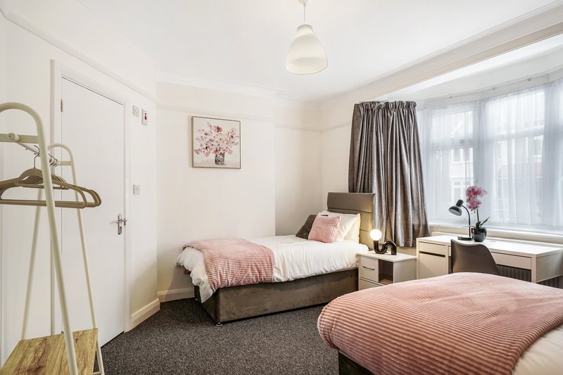 Bedroom with two single beds, pink bedding, desk, and large window with grey curtains