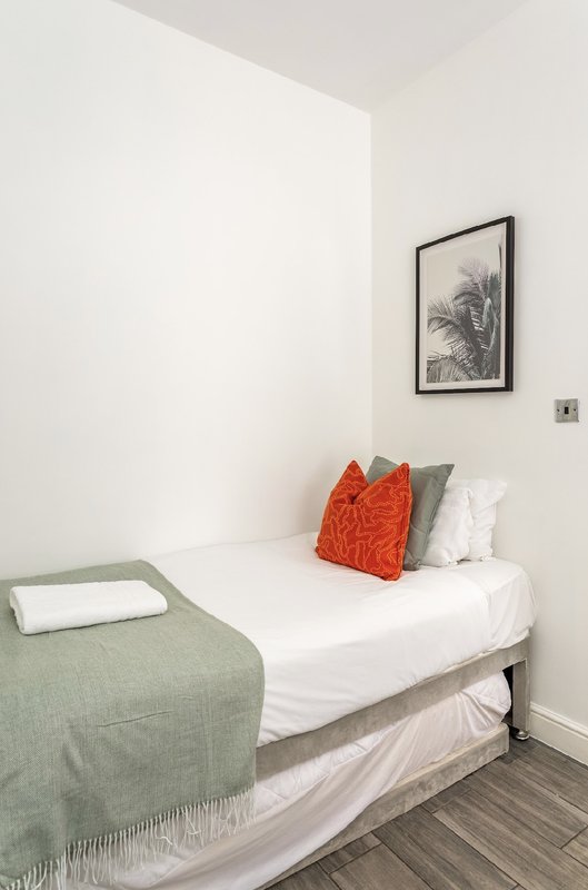 Compact bedroom with single bed, white bedding, orange and grey cushions, green throw blanket