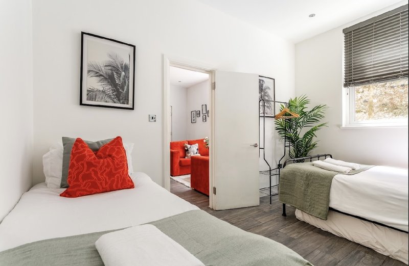 Bright bedroom with two beds, red cushion, plant, and open doorway to ensuite bathroom