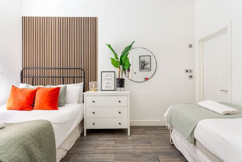 Bright bedroom with two beds, white dresser, plant, and wooden wall feature
