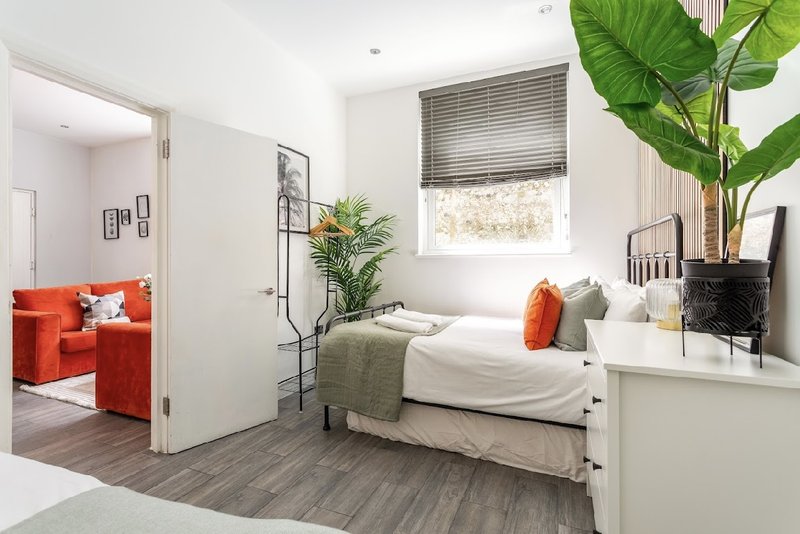 Bright bedroom with double bed, green plants, orange accent chair visible through doorway