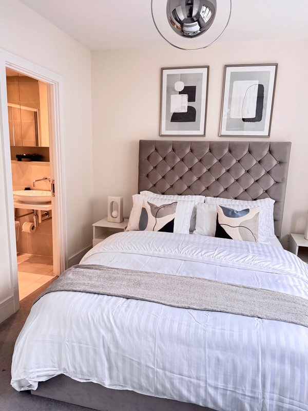 Modern bedroom with gray upholstered headboard, white bedding, and ensuite bathroom visible through open door