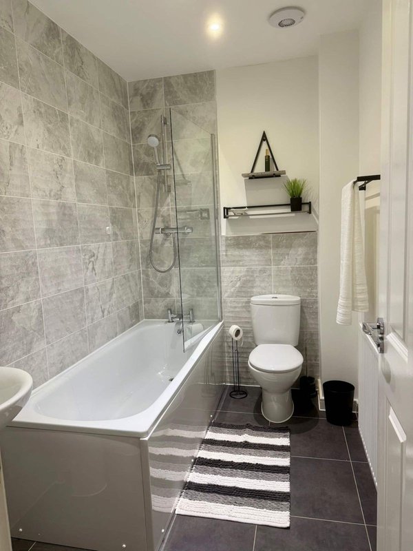Modern bathroom with white bathtub, grey tile walls, toilet, and floating shelf with accessories