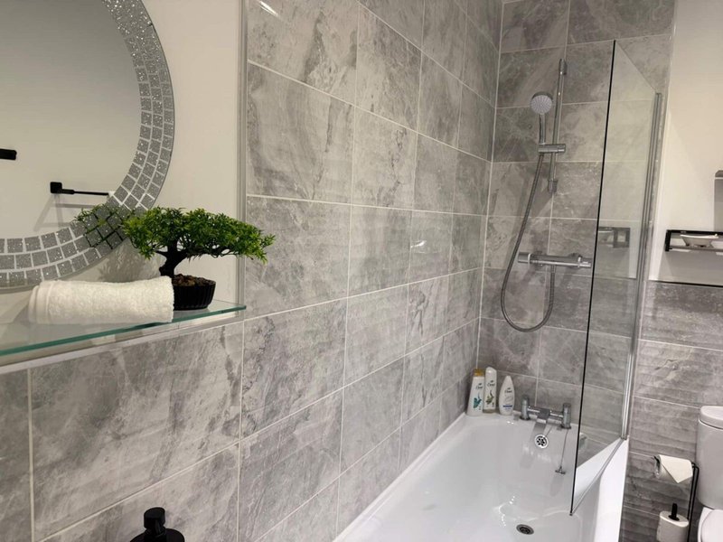 Modern bathroom with gray tile walls, glass shower enclosure, white bathtub and decorative plant on shelf