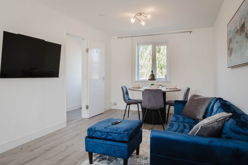 Modern living room with blue sofa, dining table, wall-mounted TV and natural light from windows